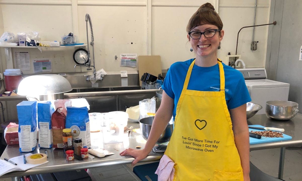 Candace Minster standing in a bright yellow apron with writing on it. She is in a commercial kitchen with bowls and baking items on the table behind her.