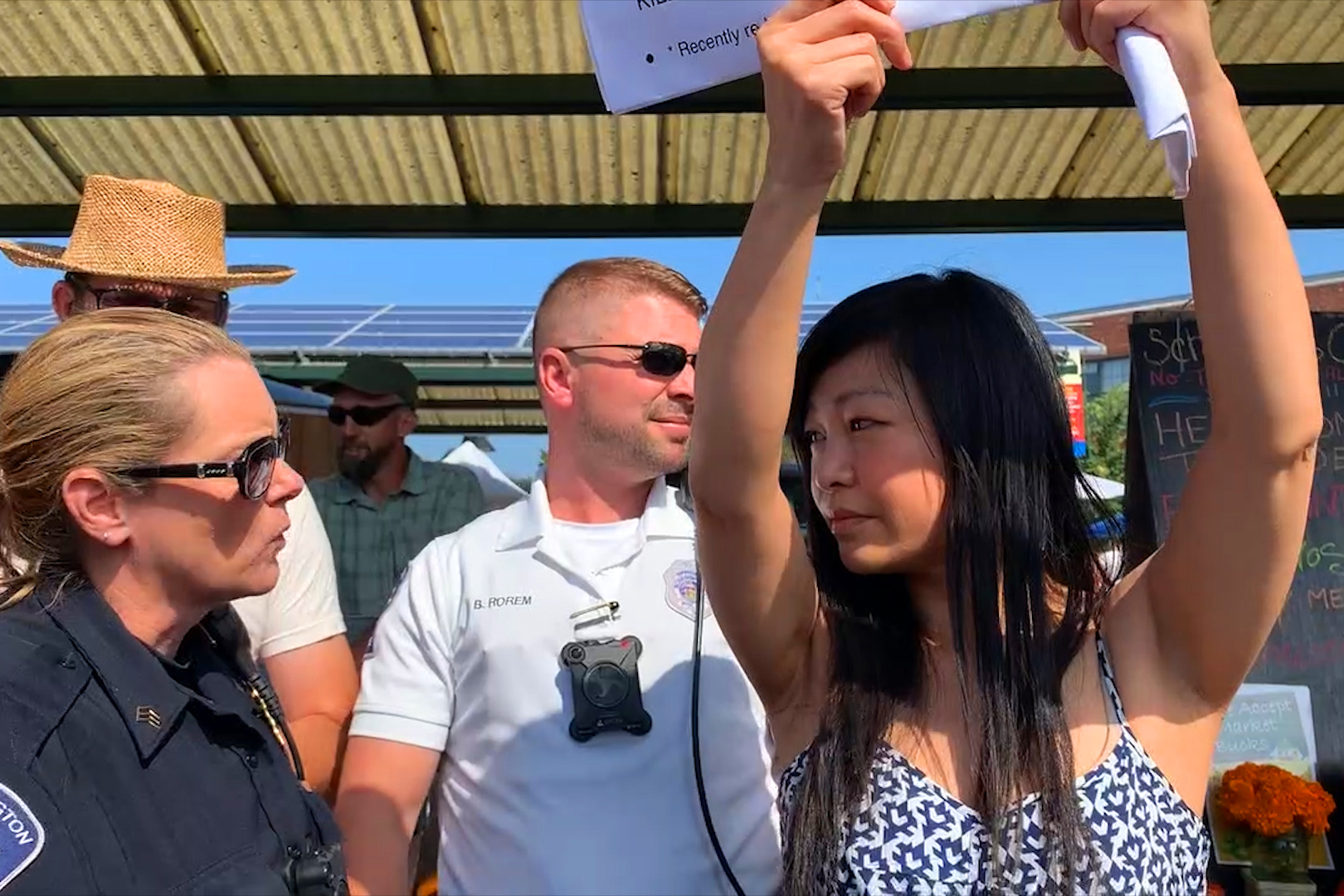 A woman in a sundress, arms holding up a sign, speaking to a female police  officer, with a male cop and a male market vendor visible behind her.