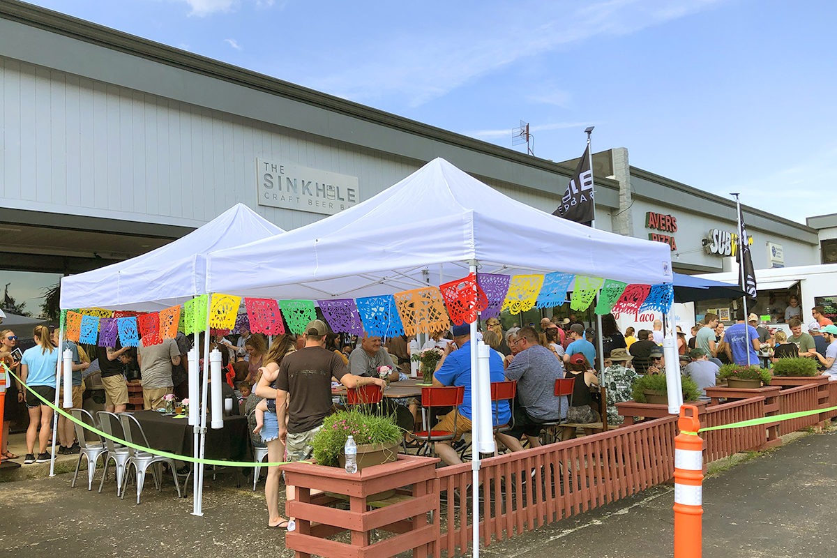 view of an outdoor seating area for food with white canopies and colorful banners. a food truck is visible in the disance and a sign on the building says "The Sinkhole Craft Beer"