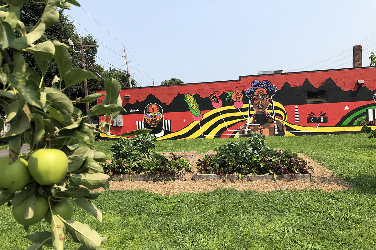 Bright mural painted onto a long building, stylized images of an african american man and woman, with red and black background and sweeping yellow and black lines. raised garden beds and apples on a tree are visible in the foreground.