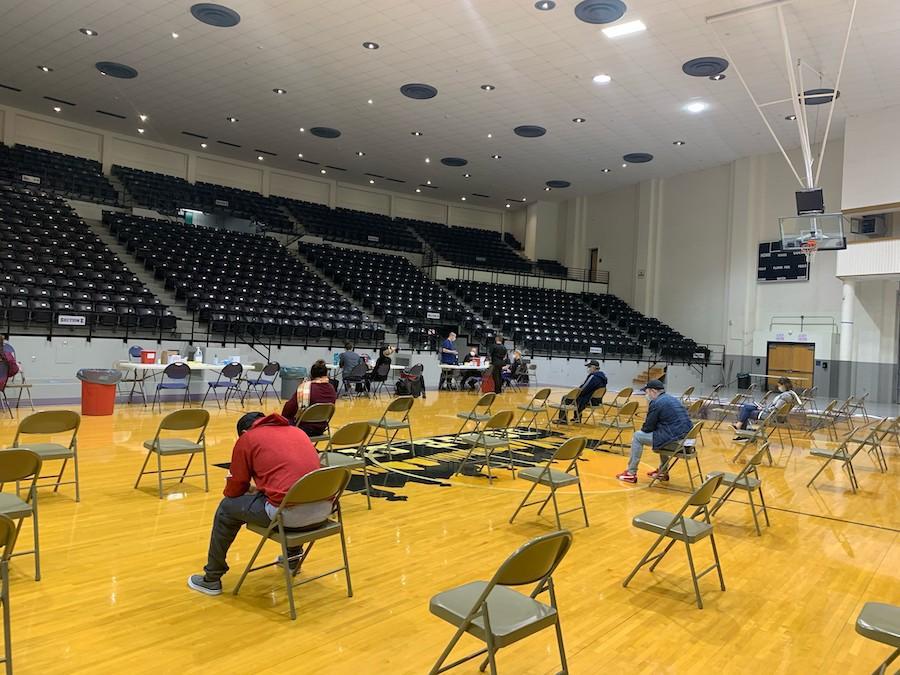 A nearly empty gymnasium with folding chairs spread out and a few people sitting in them.