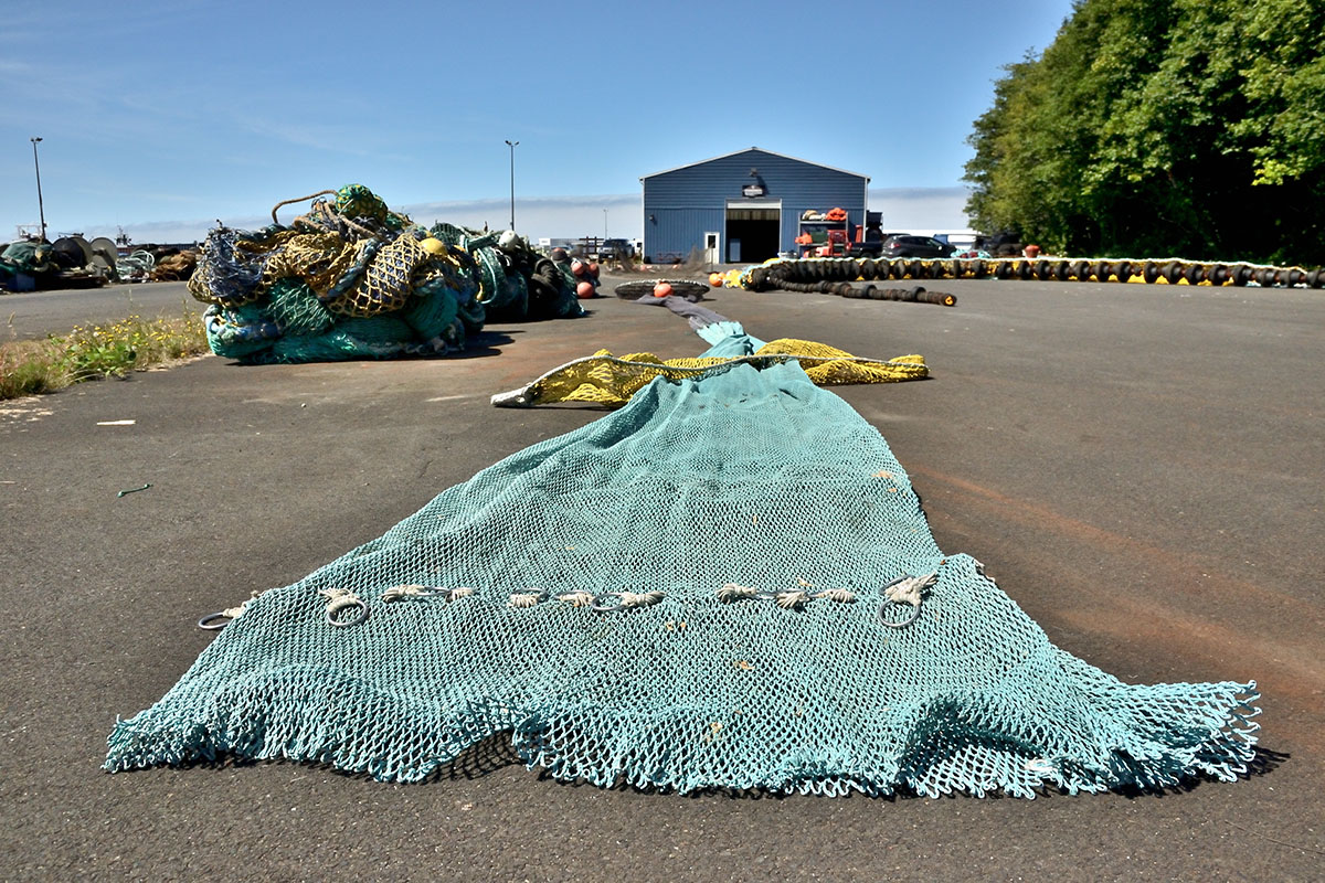 An aqua colored large commercial fishing net stretched out on asphalt where other nets are stored, a blue warehouse in the background.