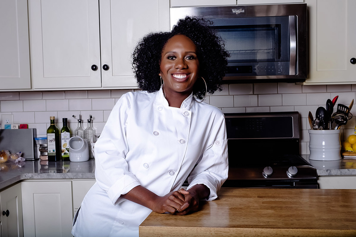 Samantha Kotey in a chef's jacket leaning on a butcher block counter top in a gleaming kitchen. 