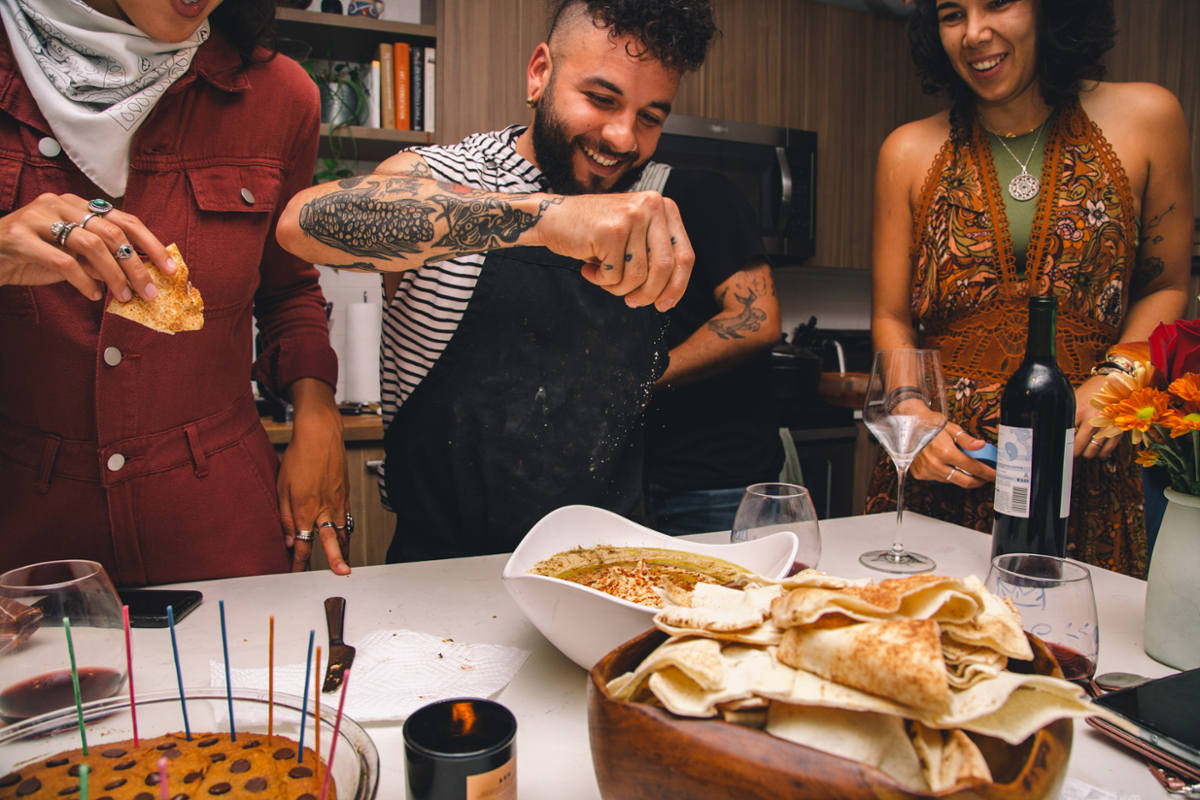 Dani Debuto, in a black apron, sprinkles salt over a dish on a table with a pile of flat bread and other food and wine. Two women on either side of him, look on, smiling.
