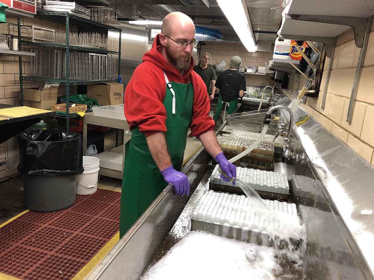 TJ Arnold stands with plastic apron at an industrial sink spraying trays of vials with water. two other staff members are seen in the background