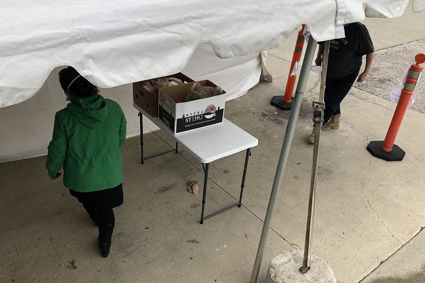 View from above of a table with boxes of food, a person with a face mask walking away while a person approachs from a space between two cones