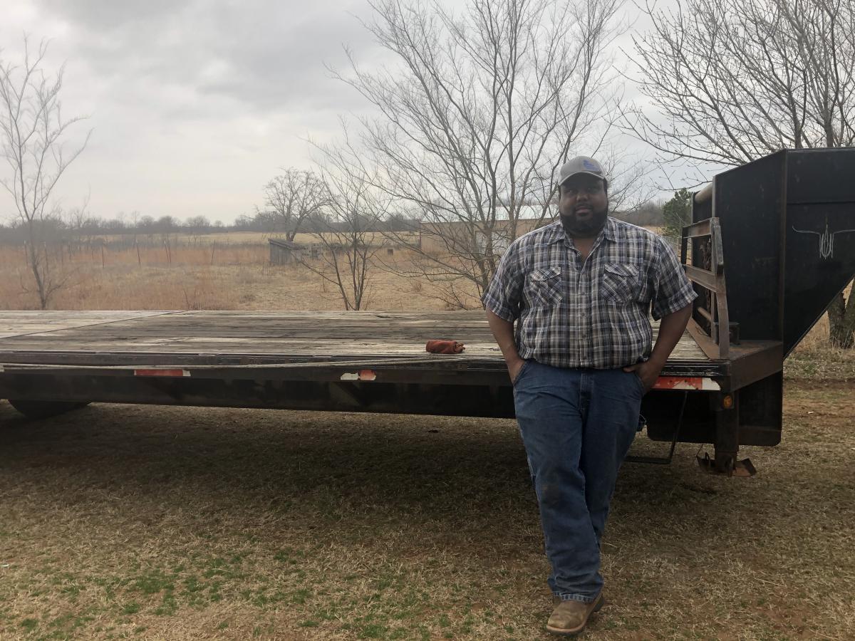 Dray Williams in plaid shirt and hat leans against a flat bed trailer with a foggy winter landscape behind him