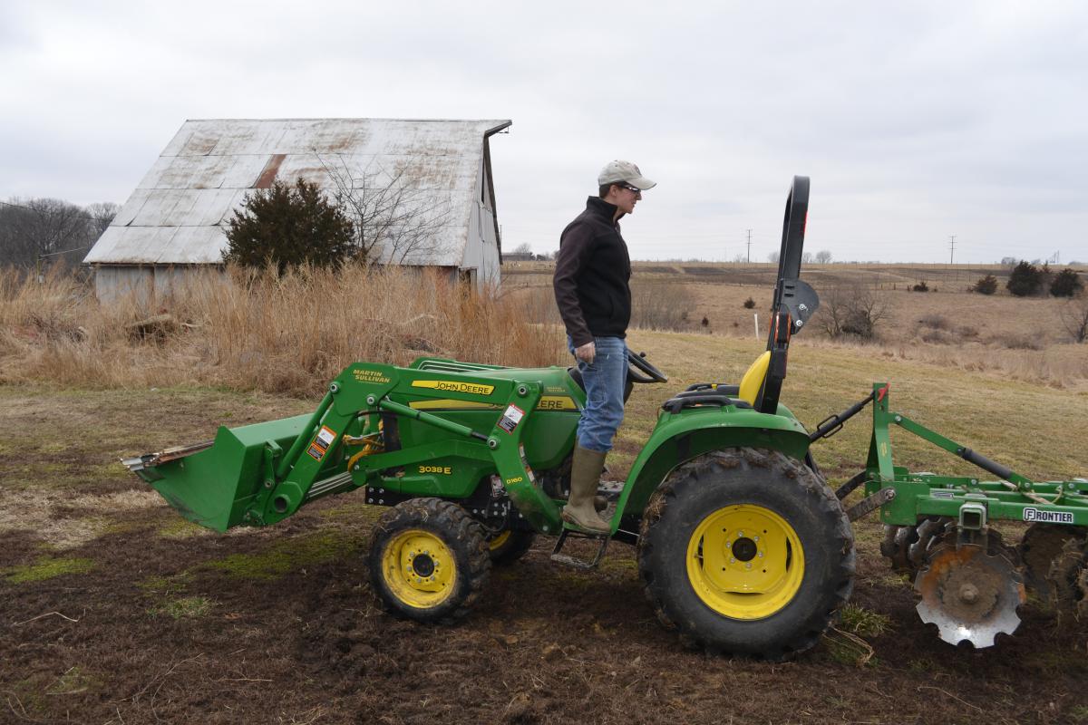 Dusty Spurgeon standing on a green tractor, facing backwards looking at the safety bar. Barn and farmland in the background.