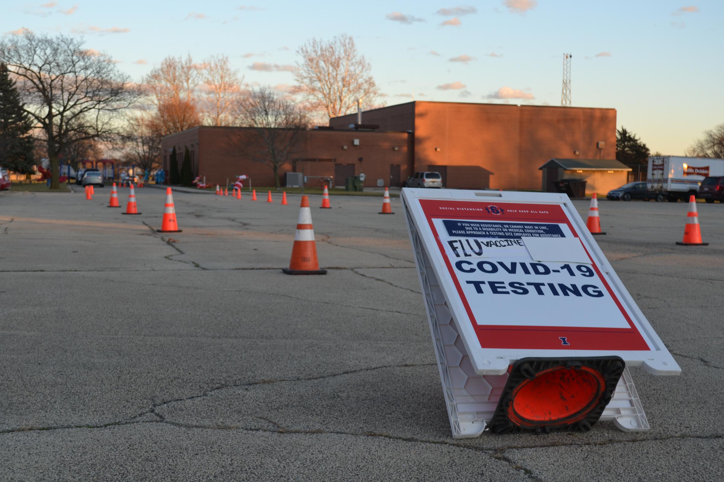 A sign on a traffic cone, tipped over says COVID Tests and Flu Shots is added in black marker. there are several traffic cones in an empty parking lot.