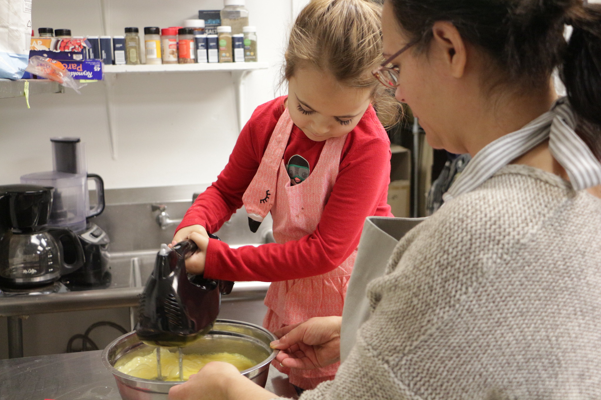 A girl in a long sleeved red shirt and a pink flamingo apron holding a hand mixer in a bowl of eggs, a woman holding the bowl, looking on.