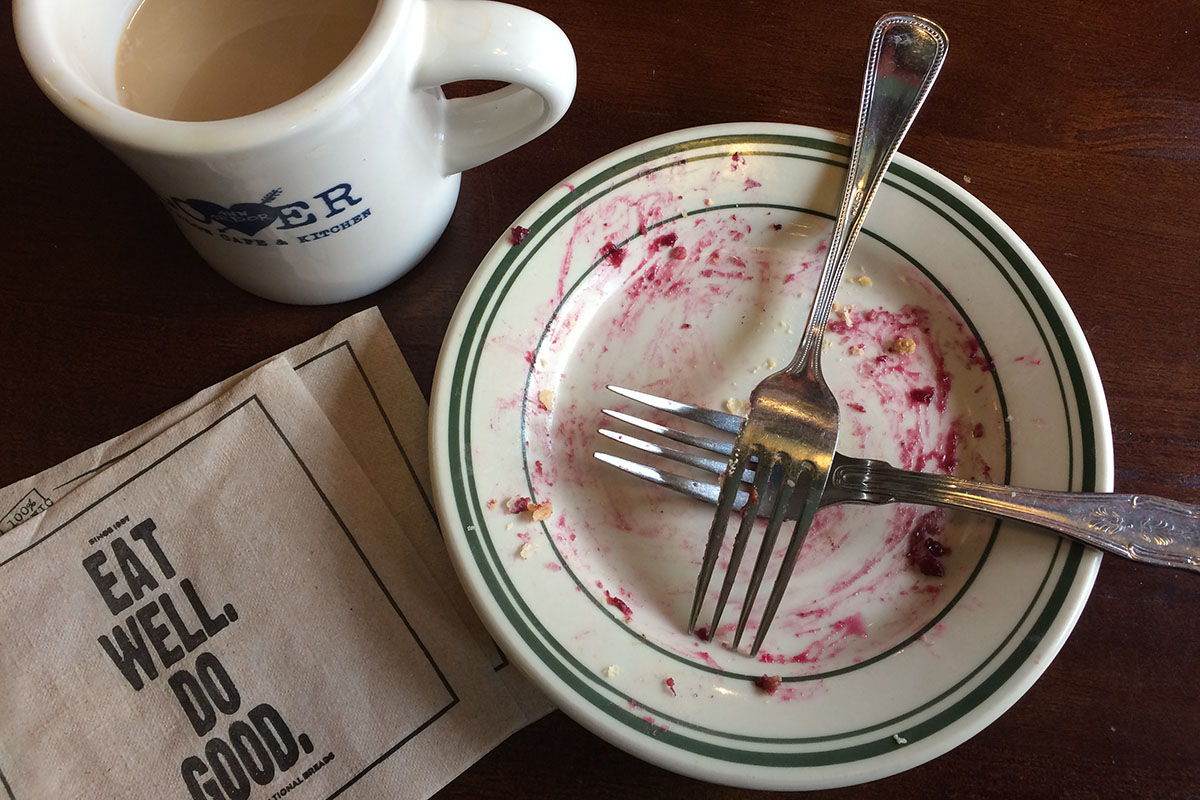 Overhead view of a small empty diner plate with two forks crossing it, and remnants of purple berry pie, next to a coffee cup and a napkin with words 'eat well, do good.'