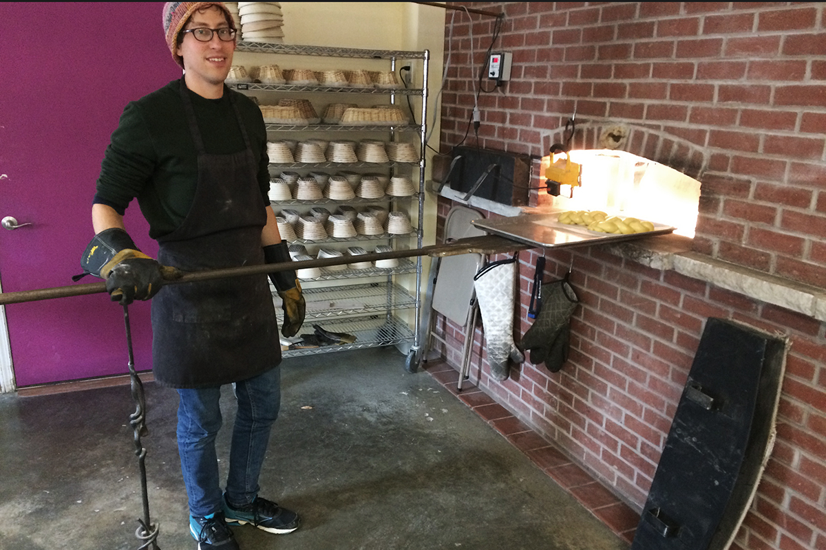 Eric Schedler standing in front of brick oven door, with long-handled pizza peel. 