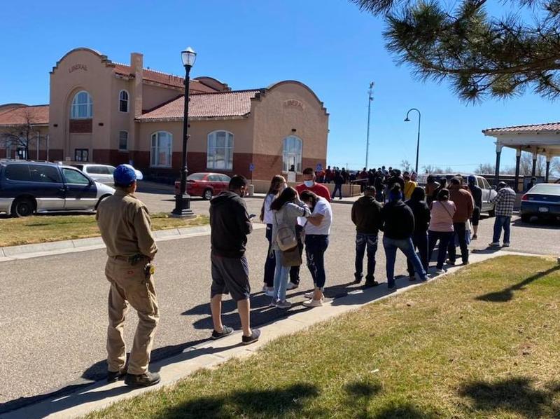 People standing in line outside, in a parking lot on a sunny day