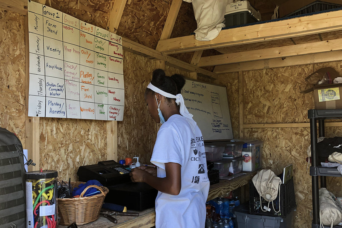 A teenage girl stands at a cash register inside of a shed/farmstand. A chart with groups and tasks is visible on the wall. 
