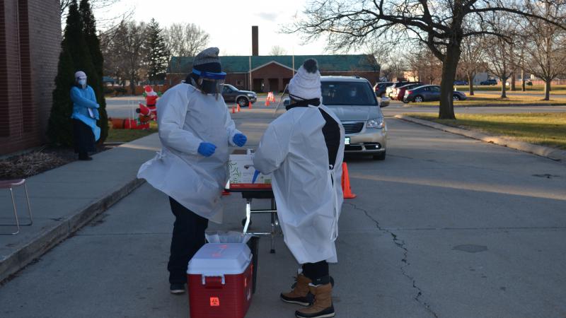 Two people in white medical protective gowns and winter hats stand at a table near a cooler outside in a parking lot as a car drives towards them