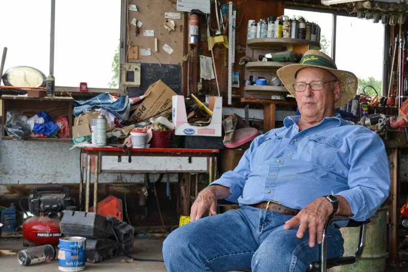 Lin Warfel wearing a straw hat and denim shirt,sitting in a chair with a cluttered garage space in the background