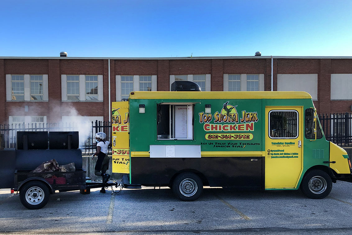 Green and yellow food truck with "top shotta jerk chicken" logo and a person stepping out the back towards a smoking barrel-style grill behind the truck. Brick building and blue sky in the background