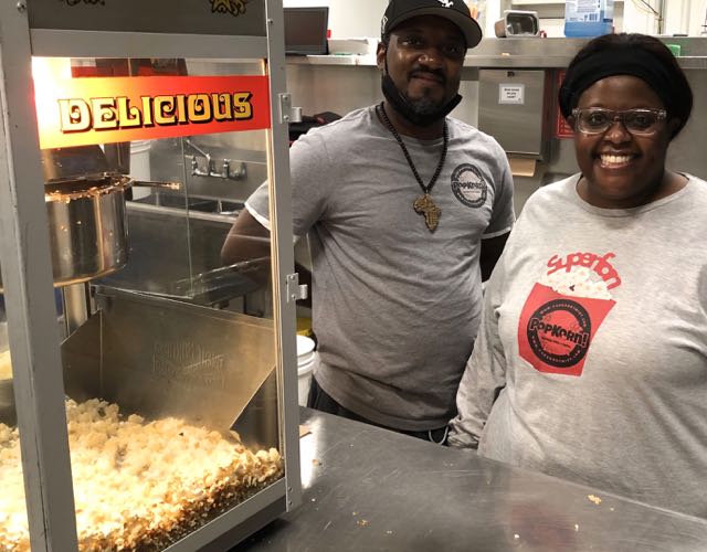 Fred Bennett and Virginia Githiri stand behind a stainless steel table in a commercial kitchen with a popcorn machine to the left. Both are wearing shirts with Popkorn logos and both are looking at the camera.