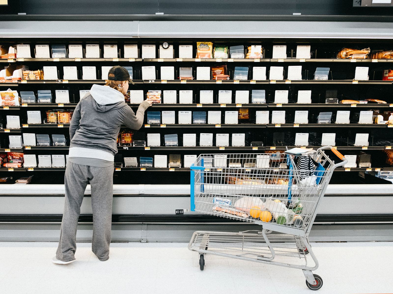 A woman in gray sweatpants and hoodie facing a nearly-empty hot dog shelf in a grocery store.