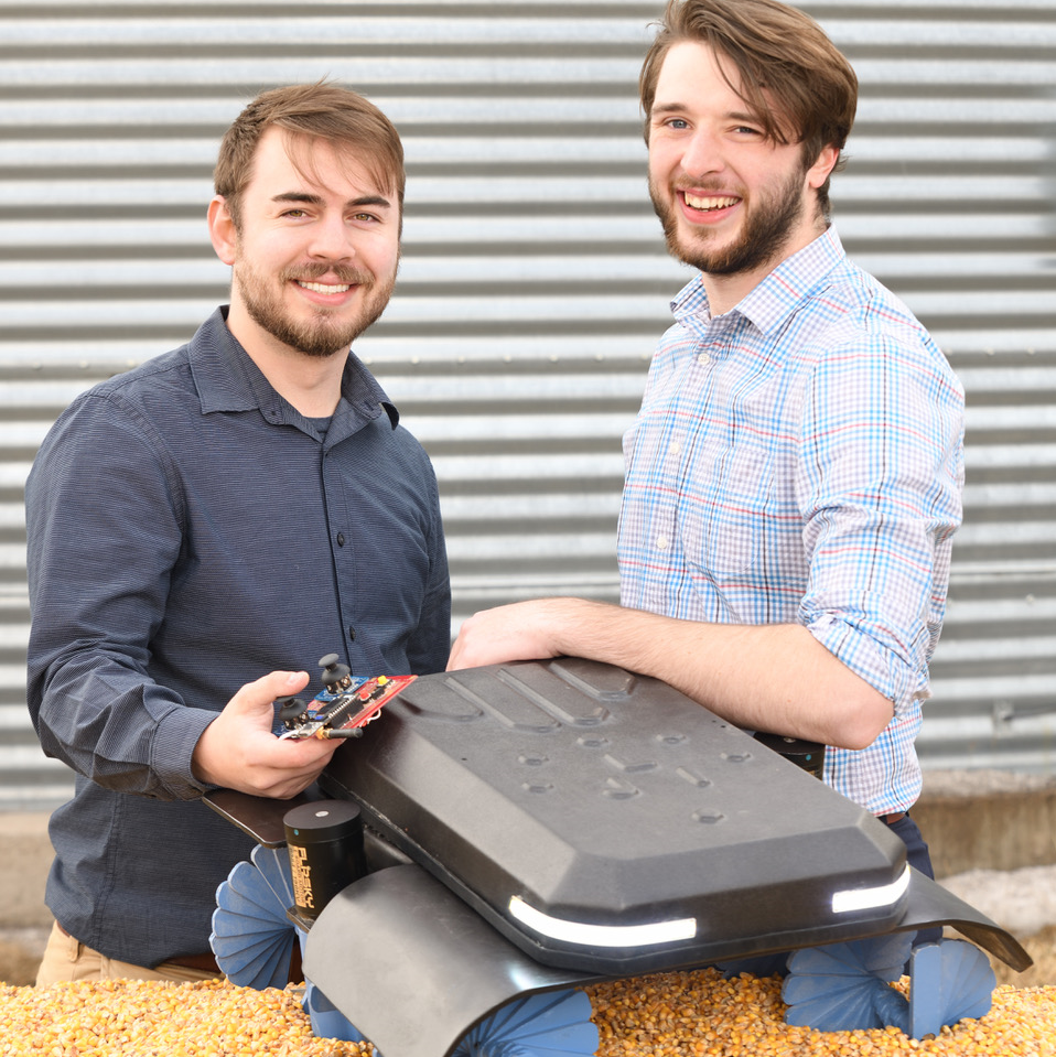 Ben Johnson and Zane Zents  smiling at camera with machine (grain weevil robot) in front of them and corregated steel background.