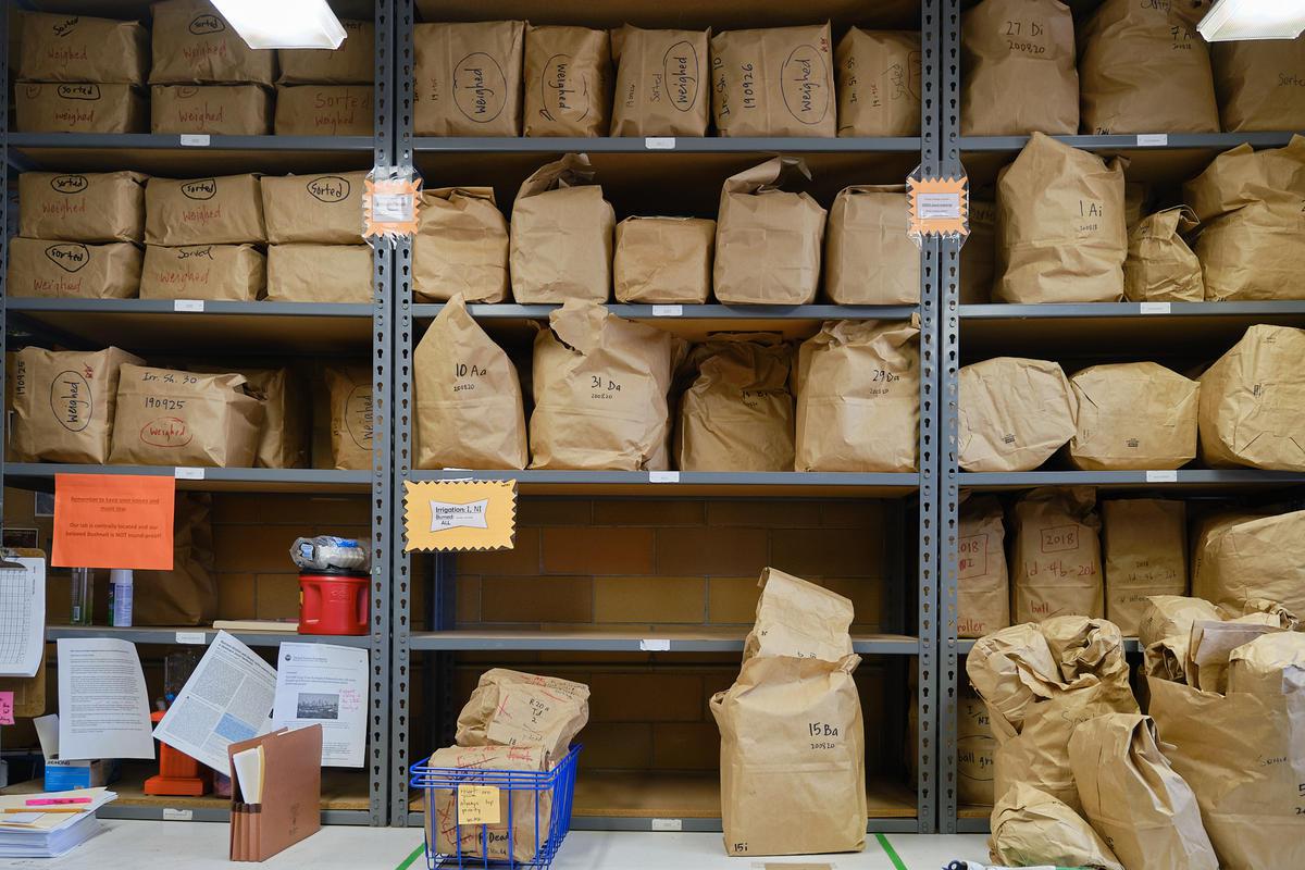 Metal shelving in a room stacked with brown paper bags, filed and folded closed, labeled with writing in black sharpie.