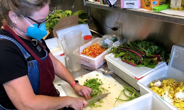 Heather Craig in apron and surgical mask chopping green vegetables in a commercial kitchen with bins of yellow mushrooms, leafy greens and chopped red and yellow peppers on the countertop