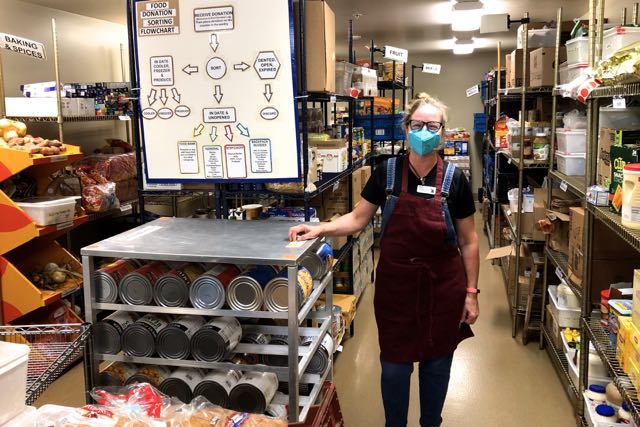 Heather Craig standing in room lined with shelves full of dry goods, with labels at top of shelves and a big diagram near where she is standing. 