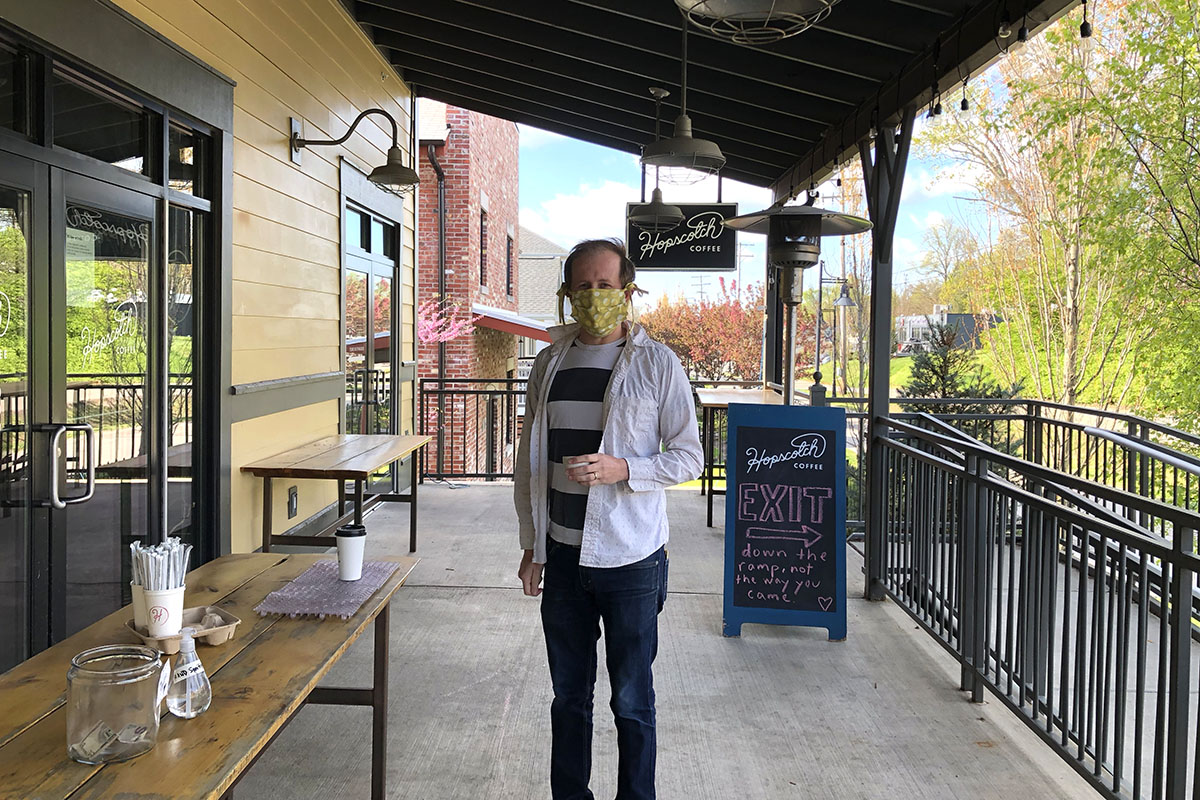 A man wearing a facemask standing on a long porch next to a table in front of a glass cafe door, Hopscotch sign in background, bright spring day. 