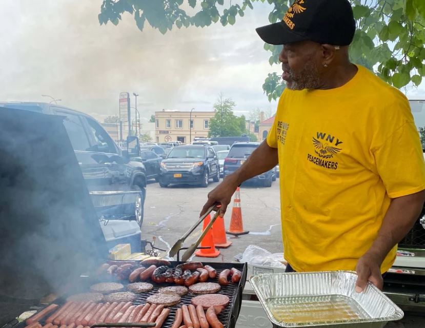 a guy in a yellow shirt at a big grill fipping hot dogs and hamburgers in a parking lot