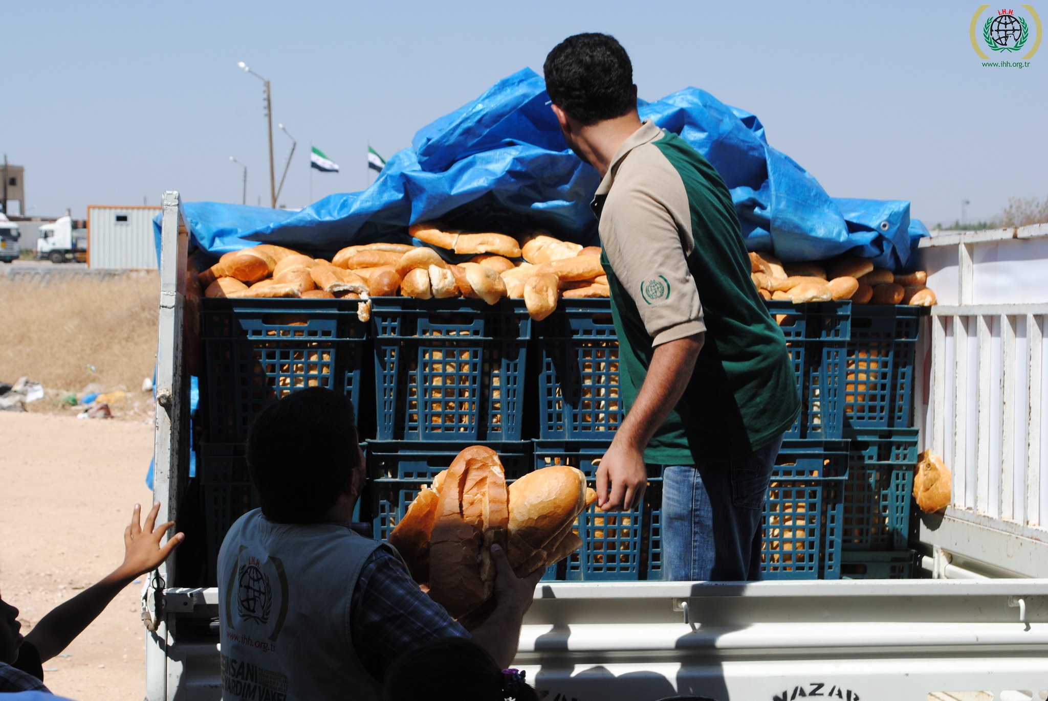 Plastic bins filled with bread on the back of a truck with a blue tarp over the top. Two people viewed from behind are unloading the bins.
