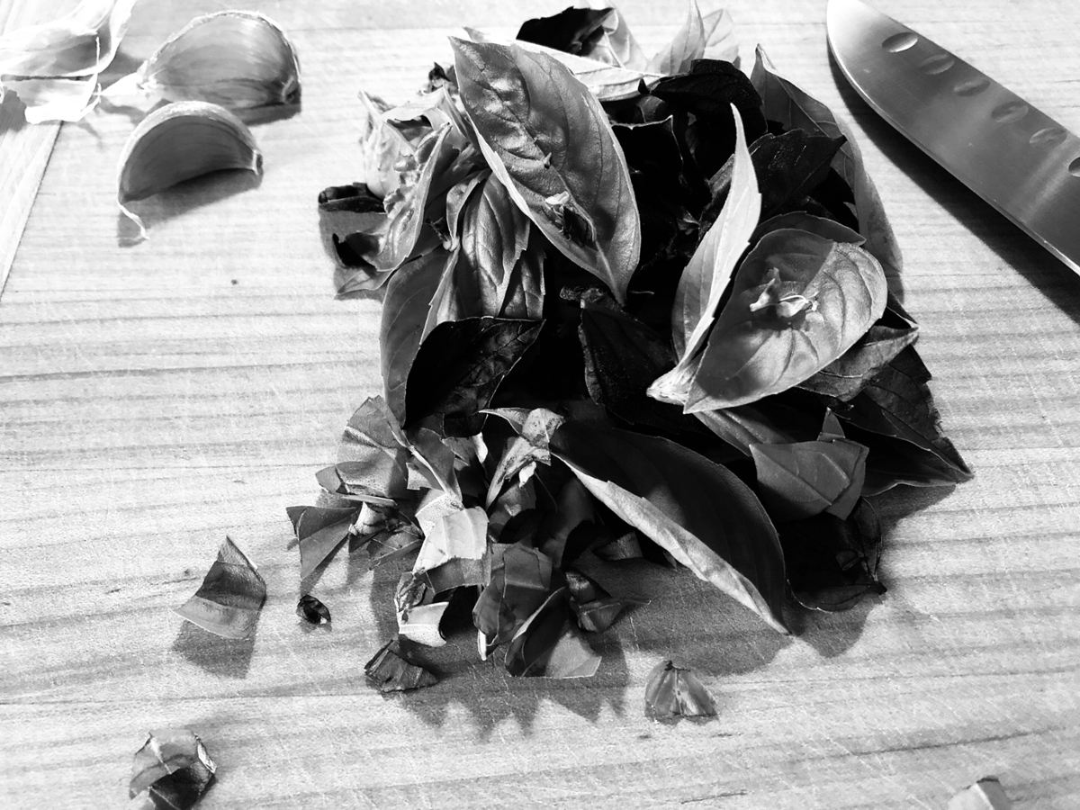 Black and white photo of basil leaves, garlic cloves and a knife blade on a cutting board