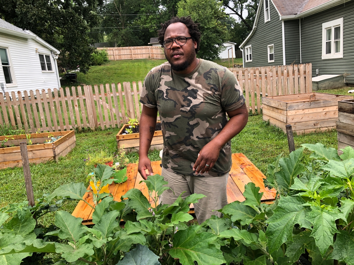 Jarrod Dortch standing behind a raised garden bed with leafy plants and a grassy yard behind him plus more wooden raised beds behind him