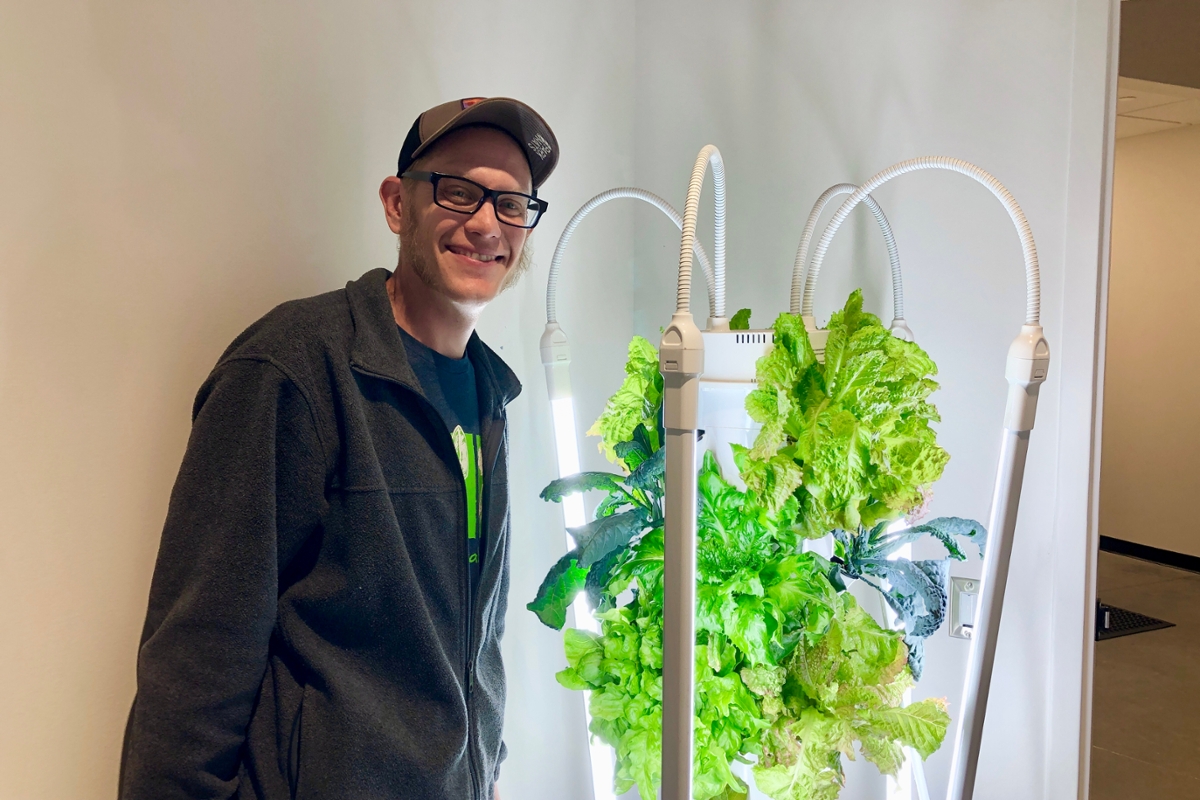 A guy in a cap and thick glasses standing next to a white tower with various leafy green an white tubes radiating out. the background is a white wall with a doorway to the right.
