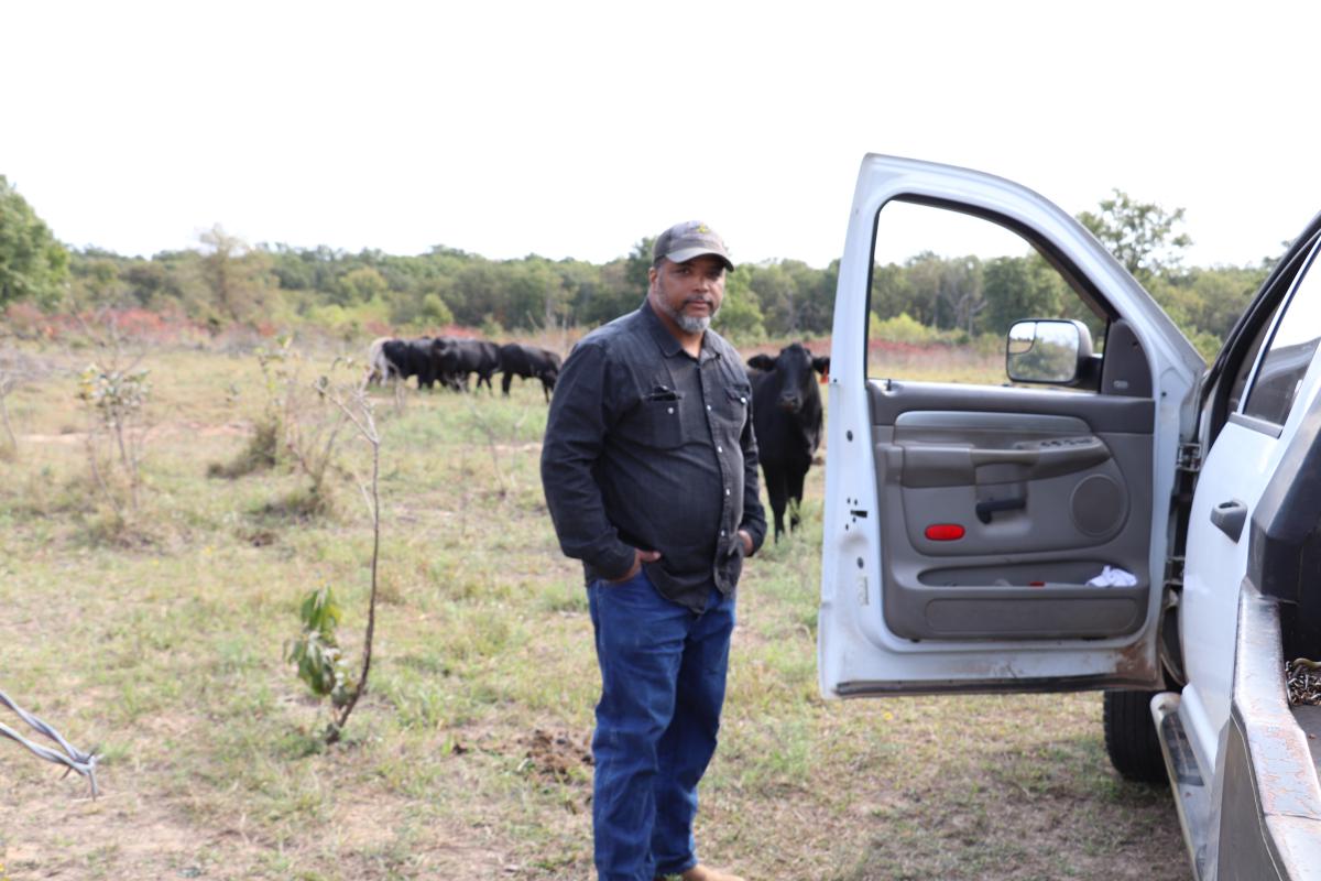 Nathan Bradford Jr. stands by an open door of a pick up, in a field with cattle in the background.