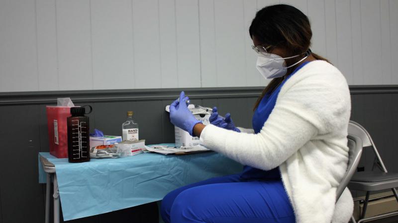 A black woman in scrubs, gloves  and face mask sitting at a table with medical supplies, handling a syringe.