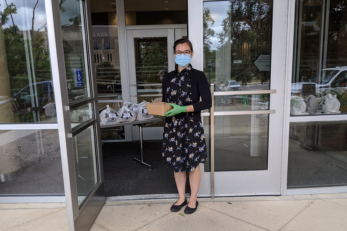 Emmie O'Connor, wearing a face mask and green gloves standing in front of glass doors with a meal box in her hand. Inside the doorway, filled plastic bags line a table. 