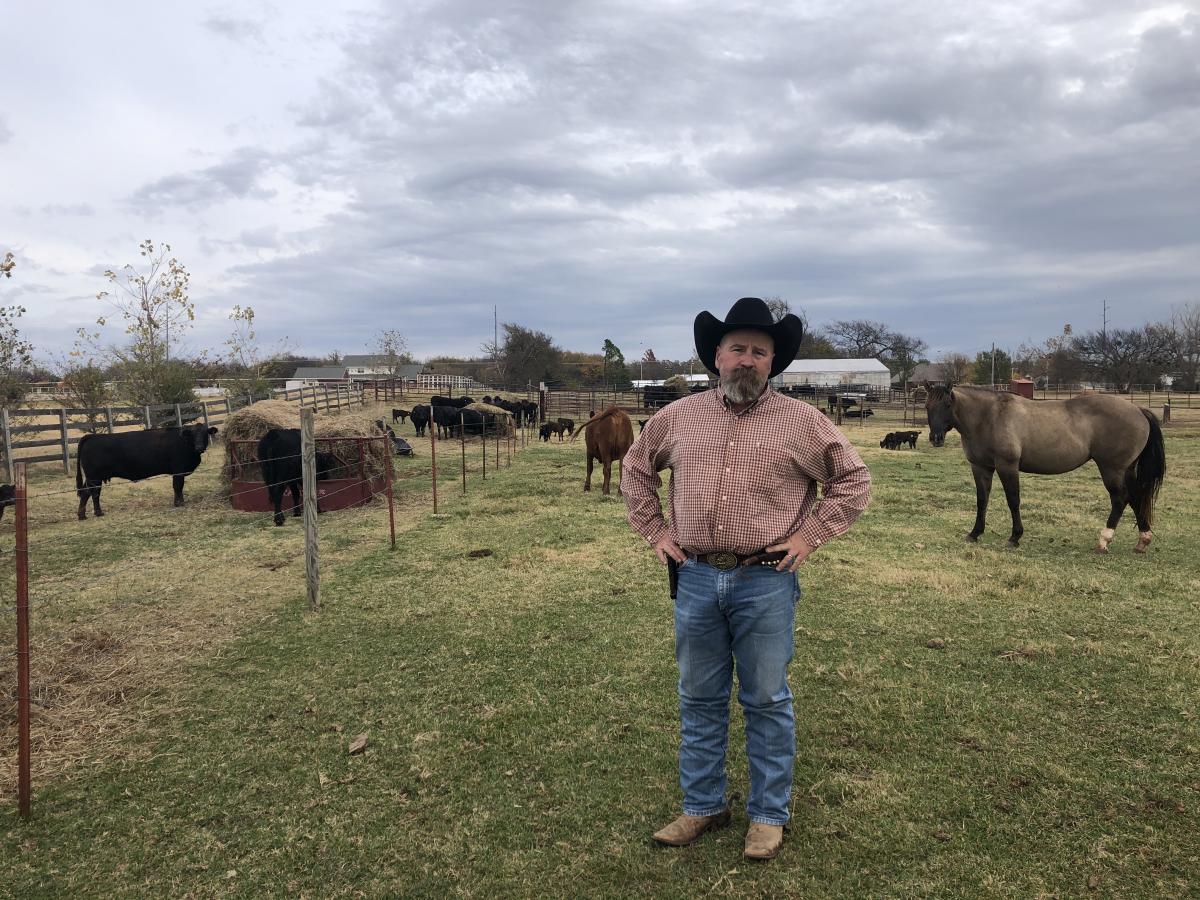 Jake Calvert stands with black cowbow hat, hands on hips, facing camera in a spacious pen with grass at his feet. In the background is a horse and several cows in various pens and an outbuilding in the distance.