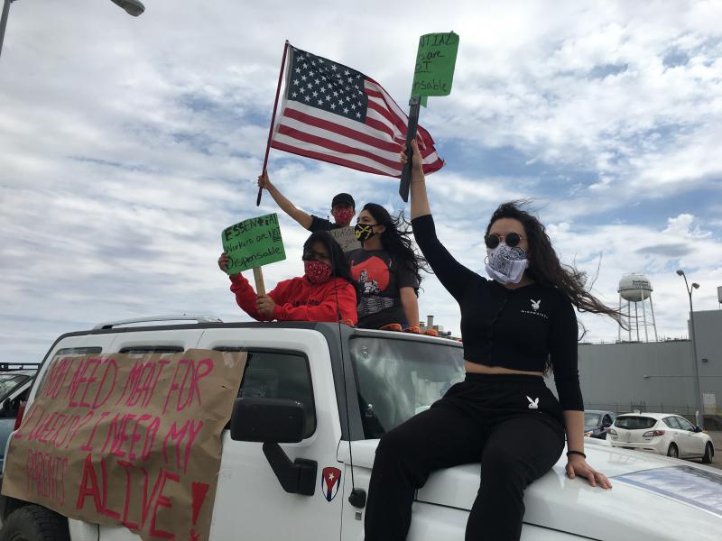Young men and women in face masks,  on a car with protest signs and American flag waving, with a factory in the background