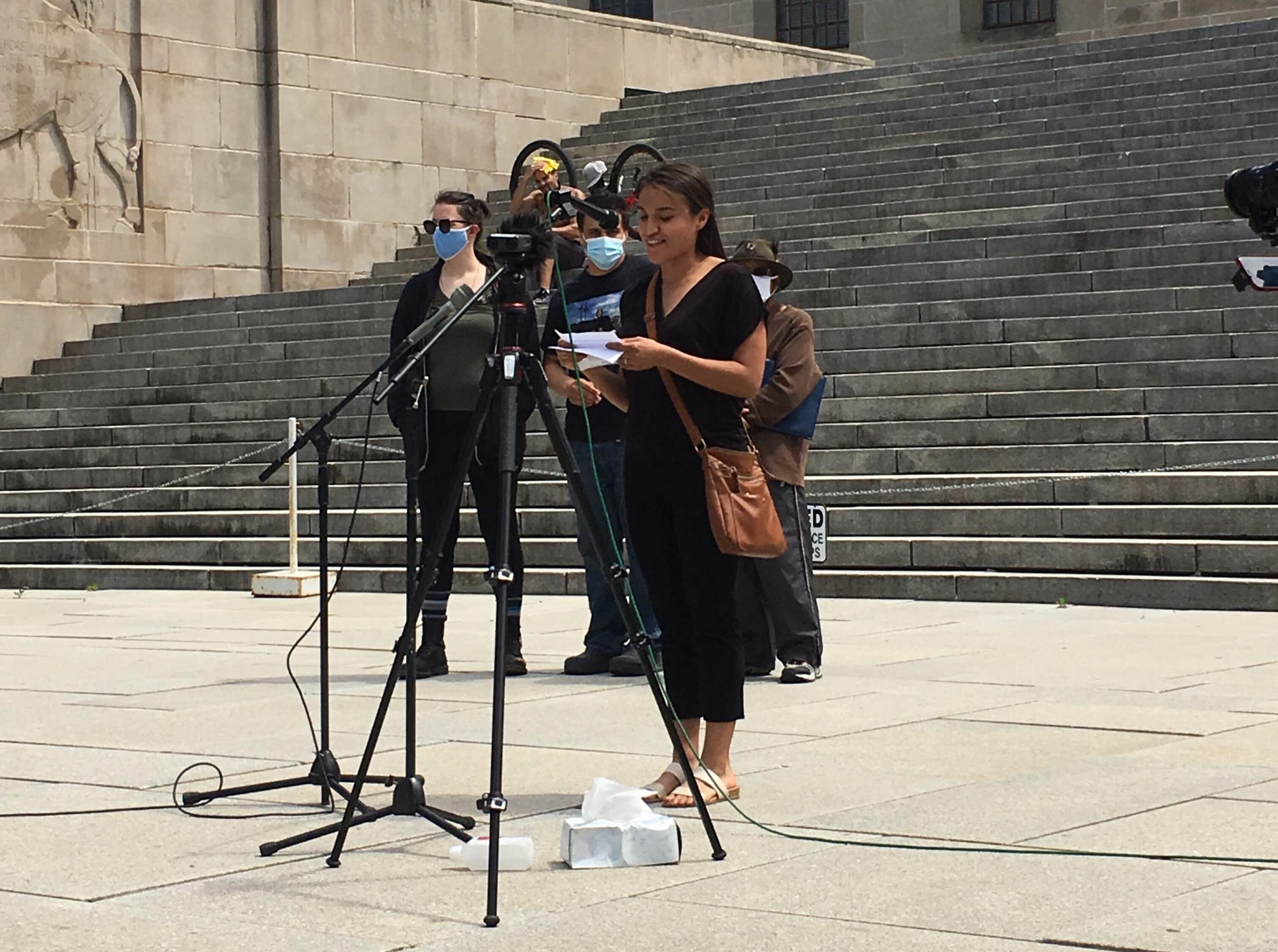 Maira Mendez speaking at a mic, with a script, people behind her, on steps, at the Nebraska State Capitol building.