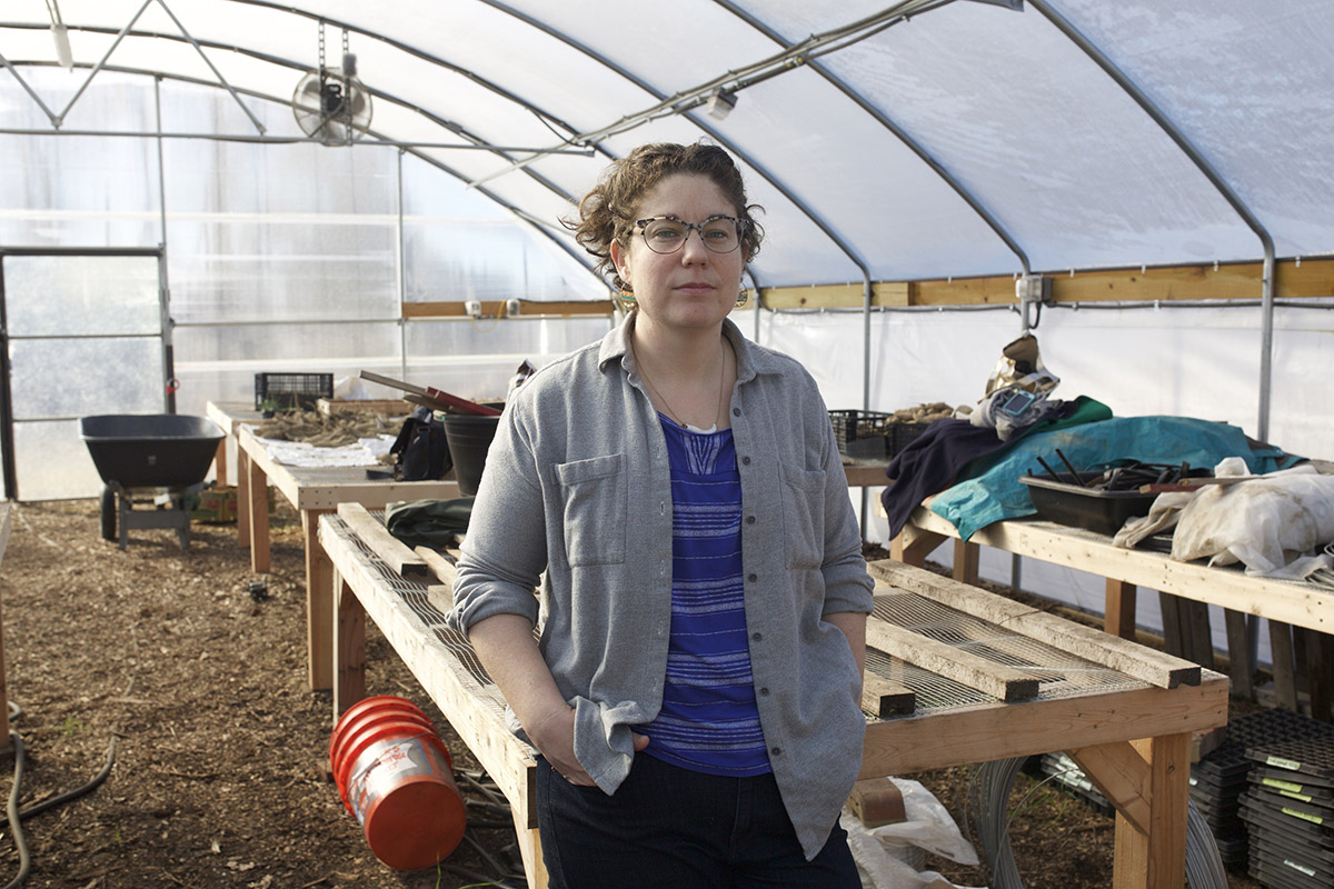 A woman standing in a mostly empty greenhouse, some potatoes and supplies in the background.