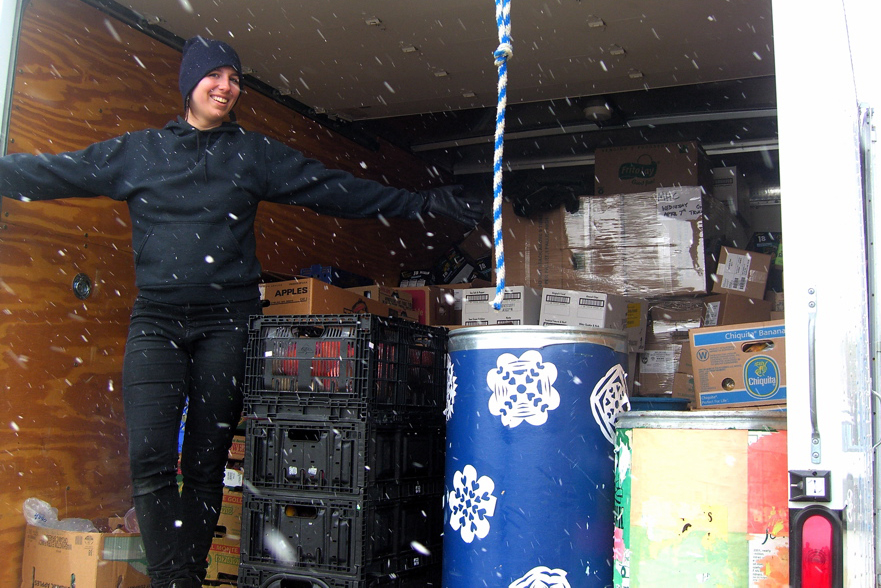 A smiling young woman stands, arms outstretched, in the back of a delivery truck, with snow falling in front of her. 