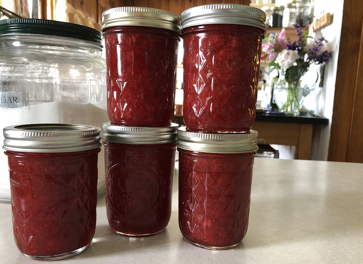 Five half pint jelly jars filled with red jam, stacked, next to a large jar with sugar inside.