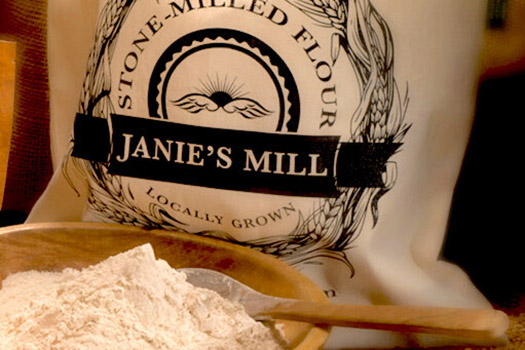 White cloth bag with Janie's Mill printed  in black, behind a bowl of flour