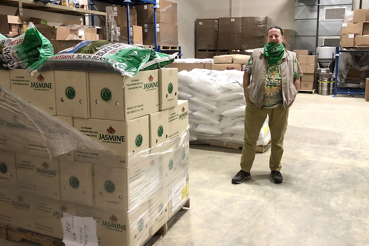 Jeff Mease with a green bandana around his chin standing in a large warehouse room next to pallets of jasmine rice and dried beans.