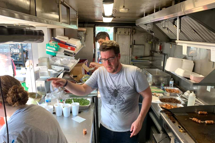 blurry shot of a man in a gray shirt with a cicada image, inside of a food truck with two other people, in action