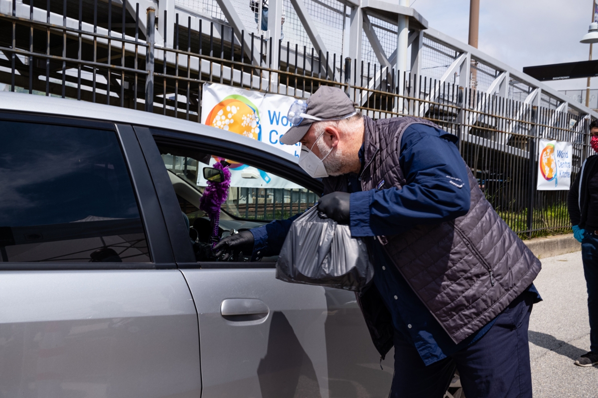 A man with a gray short beard, with face mask, gloves and hat with goggles on top, reaching into passenger side of a car, with a bag of food.