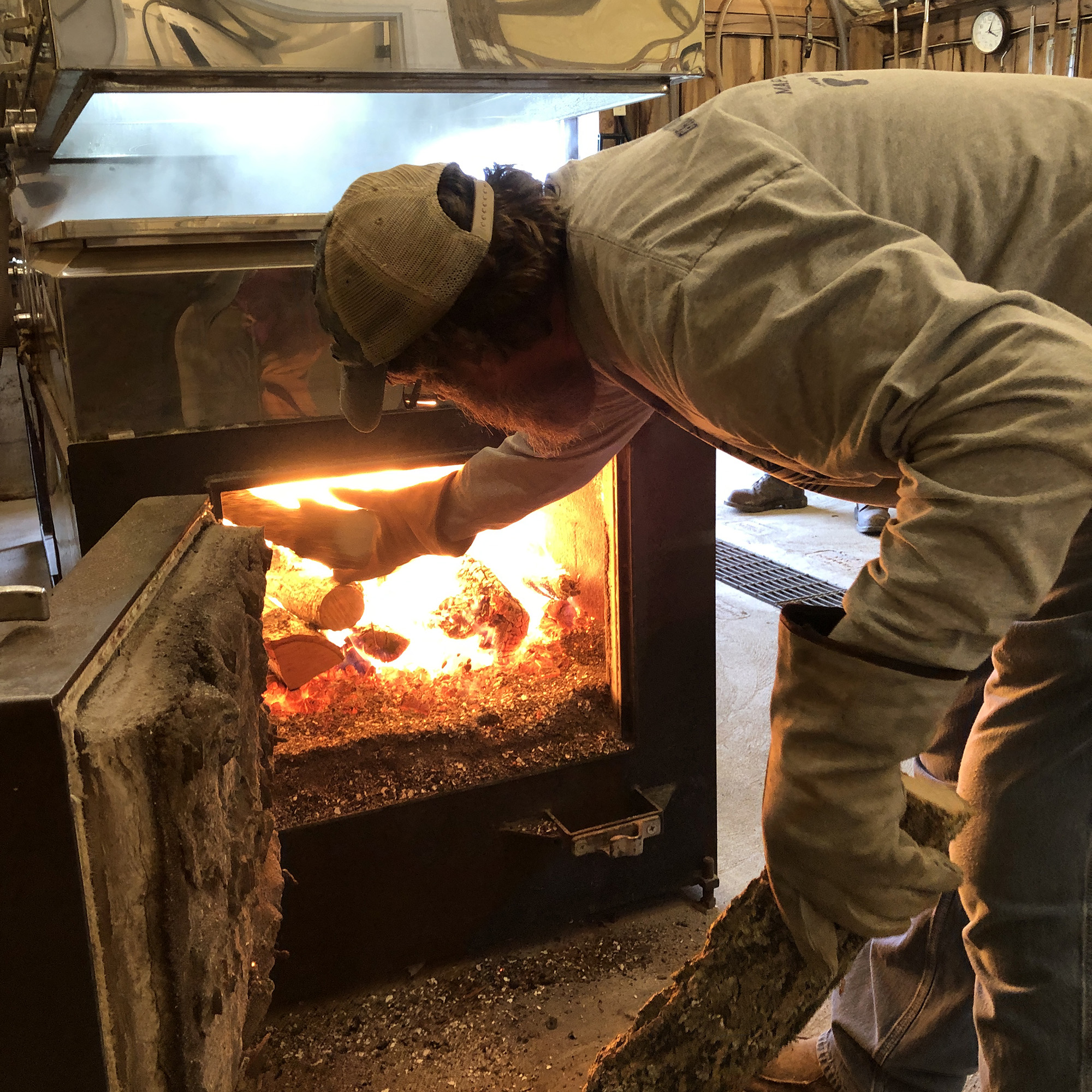 Bearded man with ball cap and leather gloves loading logs into an oven or wood stove