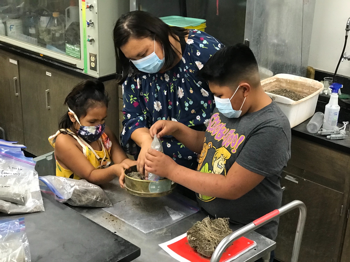 adult and two children wearing face coverings, looking down and working with a round metal tray and ziplock bags in a lab setting