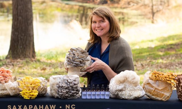 Laura Stewart standing behind a table outdoors, the table is a display of various mushrooms growing out of light colored blocks