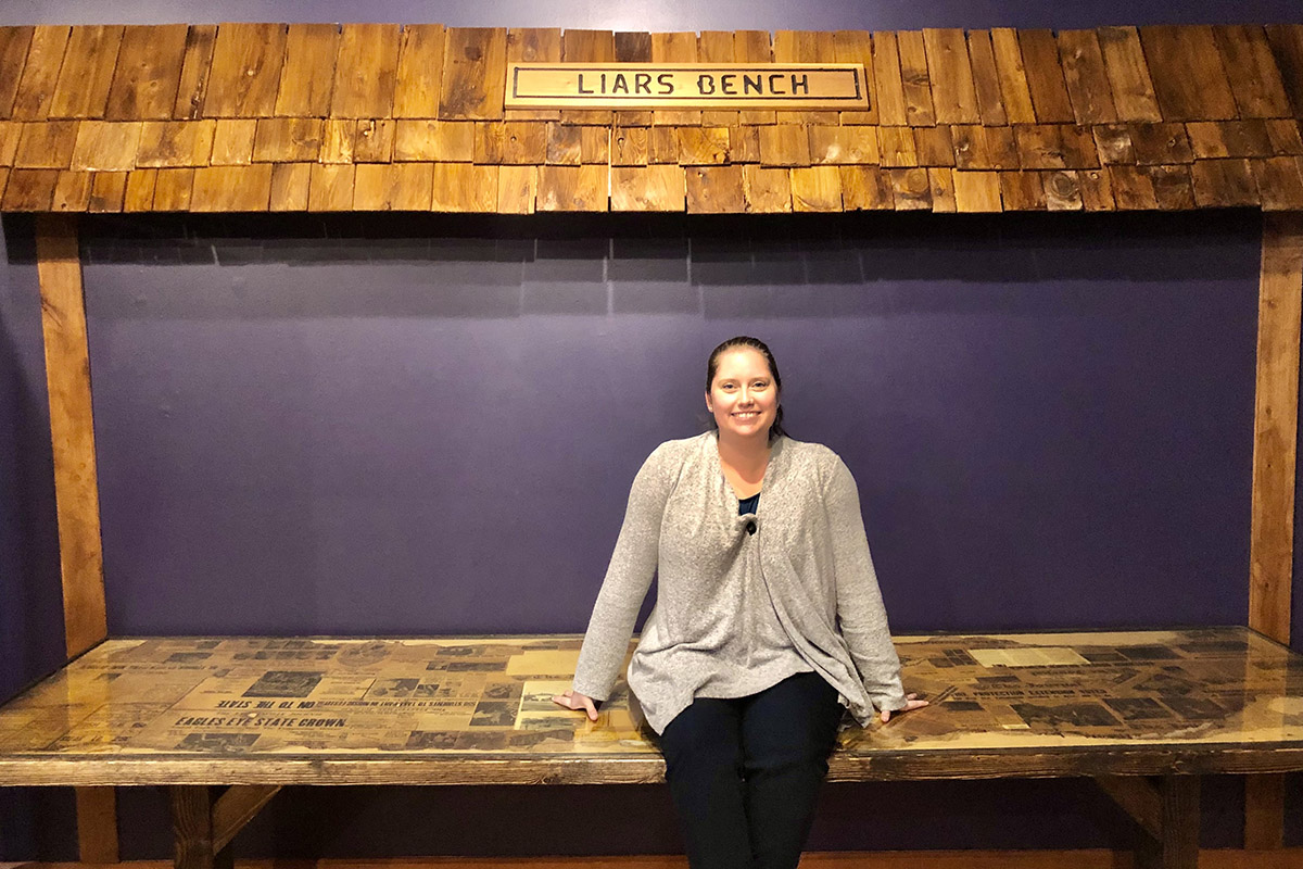 Hilary Fleck siting on a table with purple background and a shingled roof over the table with a sign that reads 'liars bench'